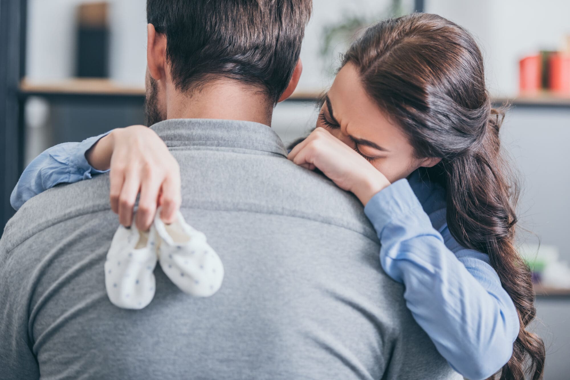 woman crying, hugging man and holding baby socks at home, grieving disorder concept Baby Loss