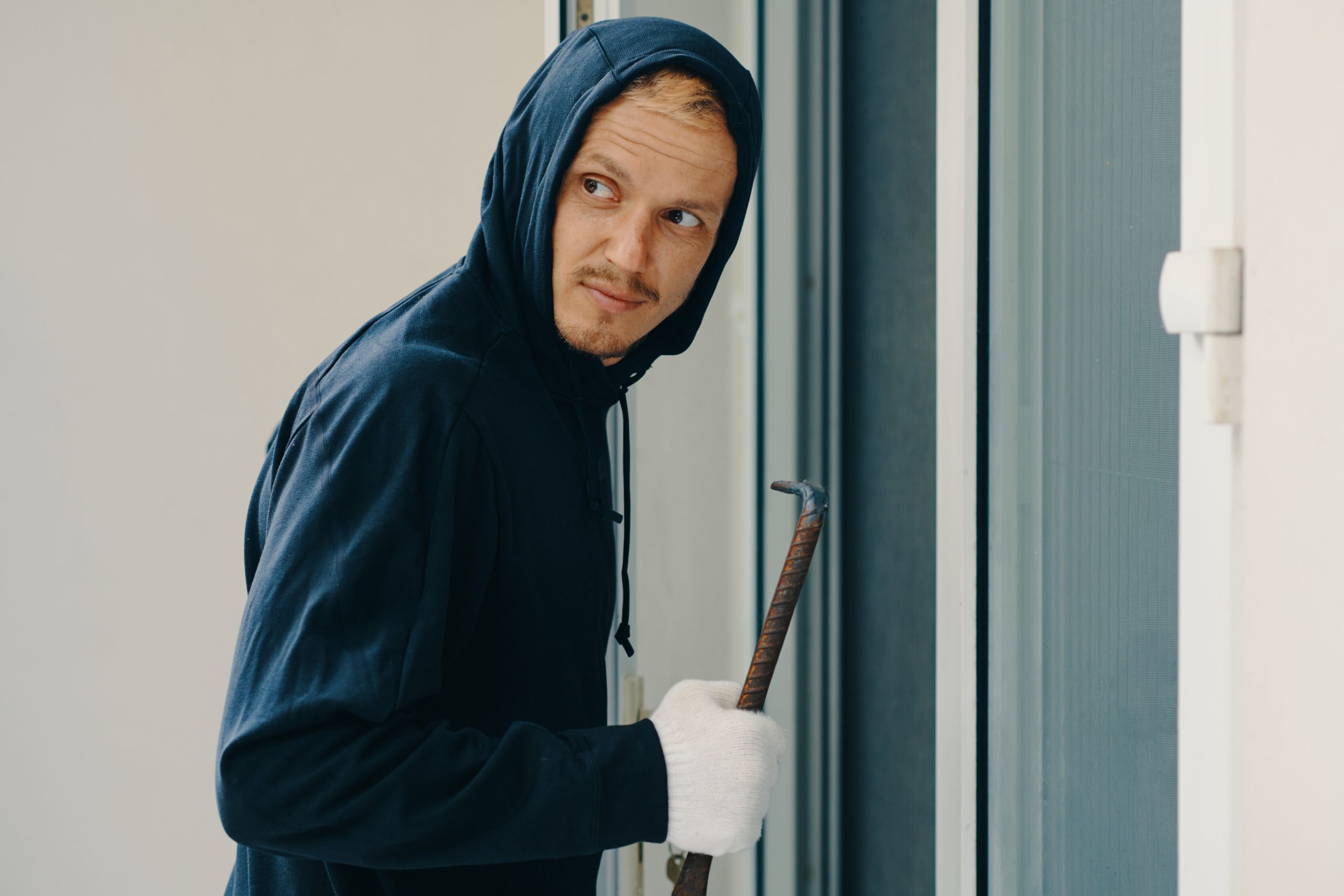 Man in a navy hoodie and white gloves gripping a crowbar at a door, looking to the side.