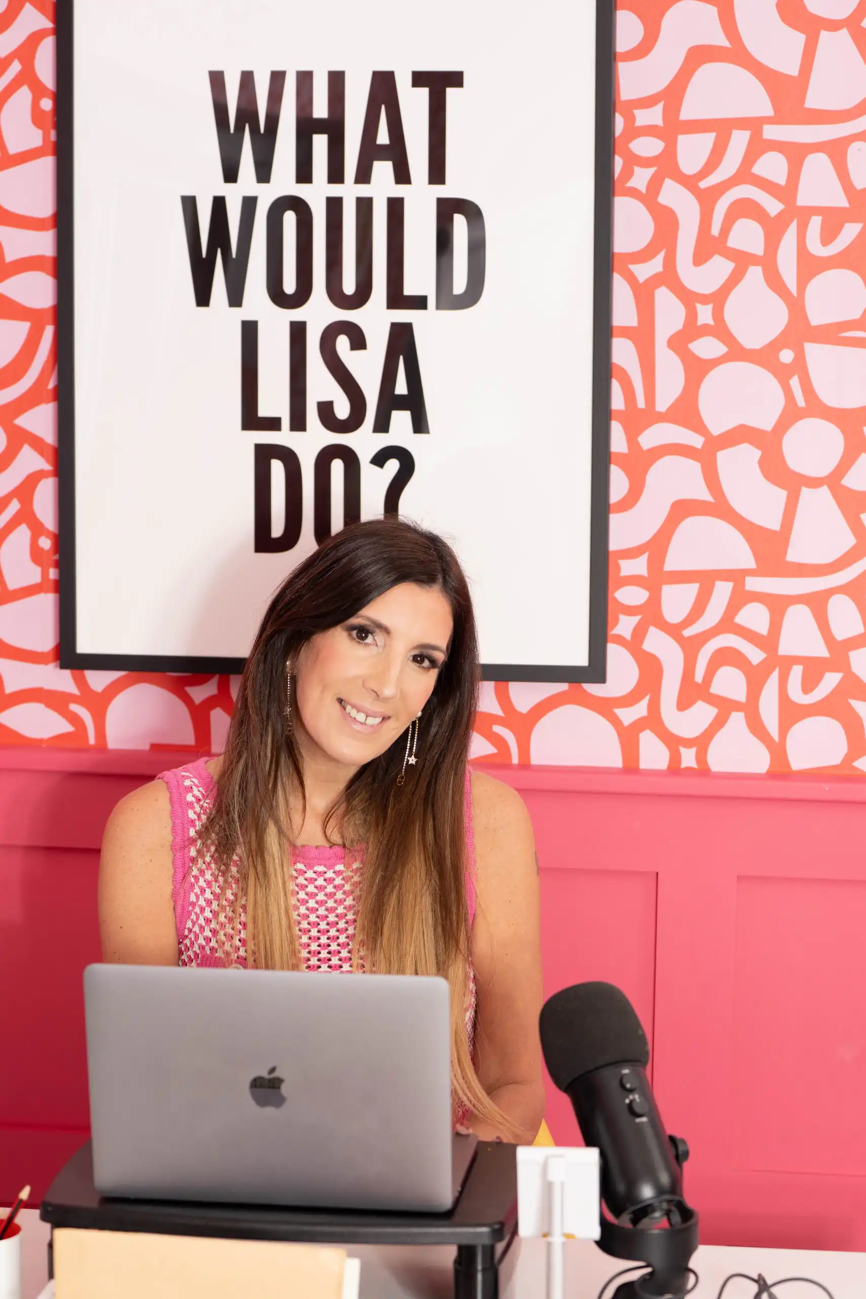 SmilingLisa Johnson at a pink desk with a laptop and a microphone; poster behind reads 'WHAT WOULD LISA DO?'