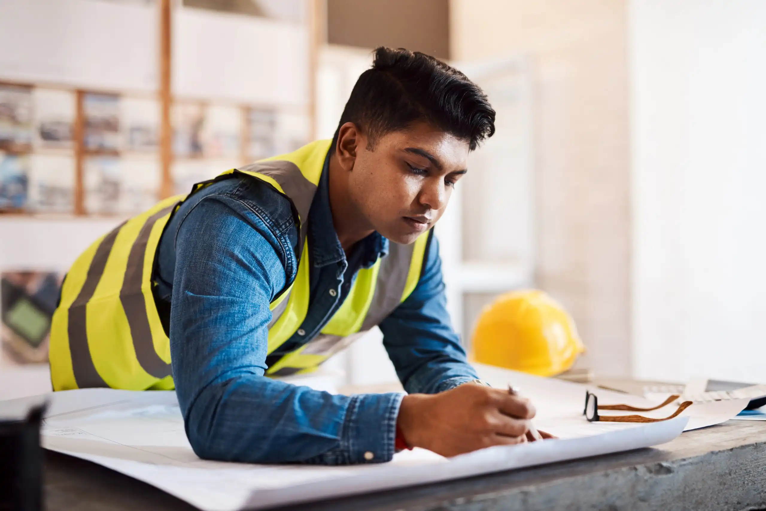 Man in a yellow safety vest and denim shirt writes on large blueprints at a worktable in a bright workshop after getting his job back in construction with a yellow hard hat in the background.
