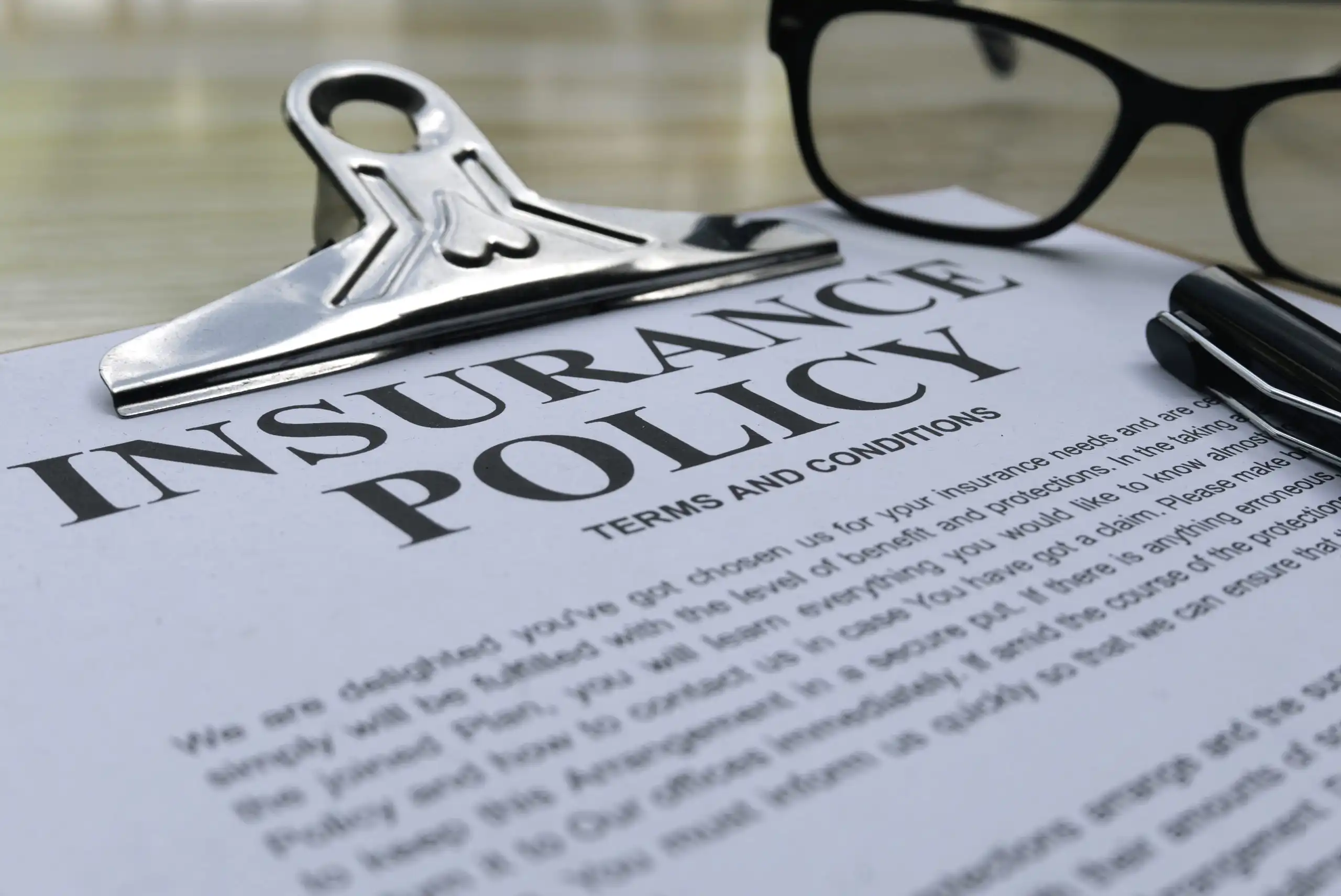 Close-up of an insurance policy document with a metal clipboard and black-framed glasses resting on it on a desk