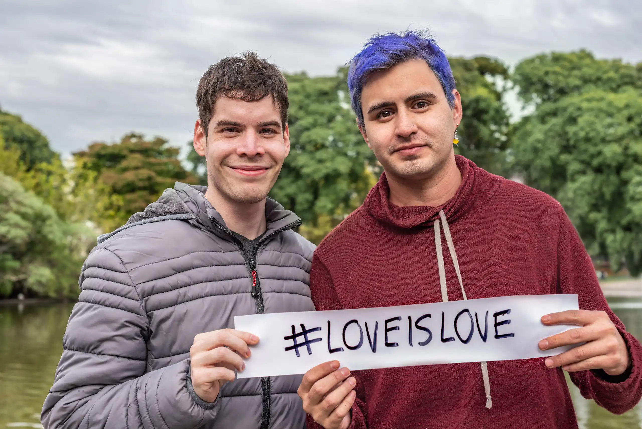 Two men smiling outdoors while holding a sign that reads #LOVEISLOVE.