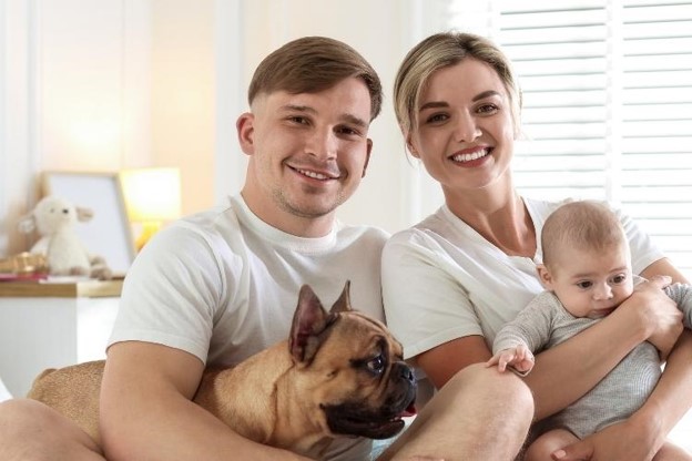 A smiling couple holding their baby and sitting next to a French Bulldog puppy