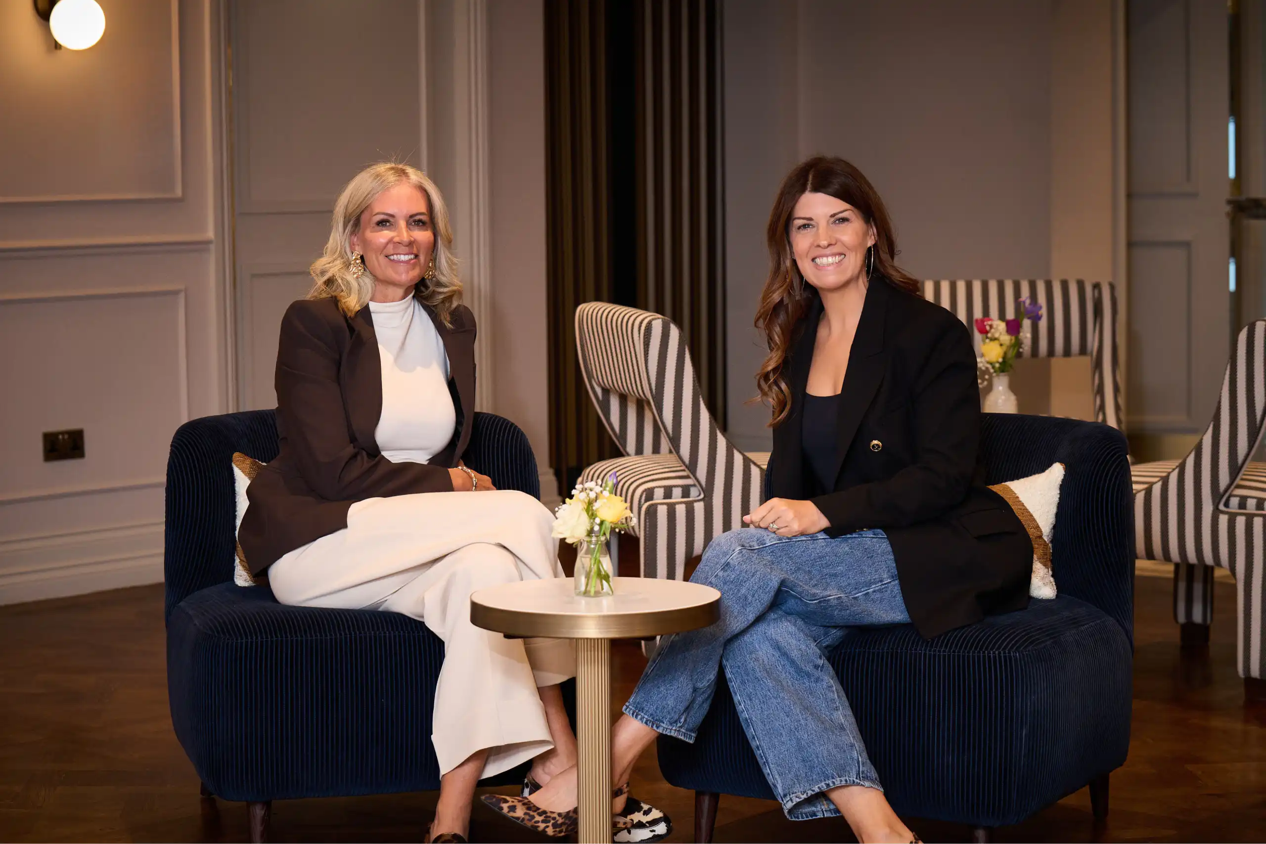 Two women sit on blue velvet chairs in a chic lounge, smiling at the camera with a small flower arrangement on the table between them.