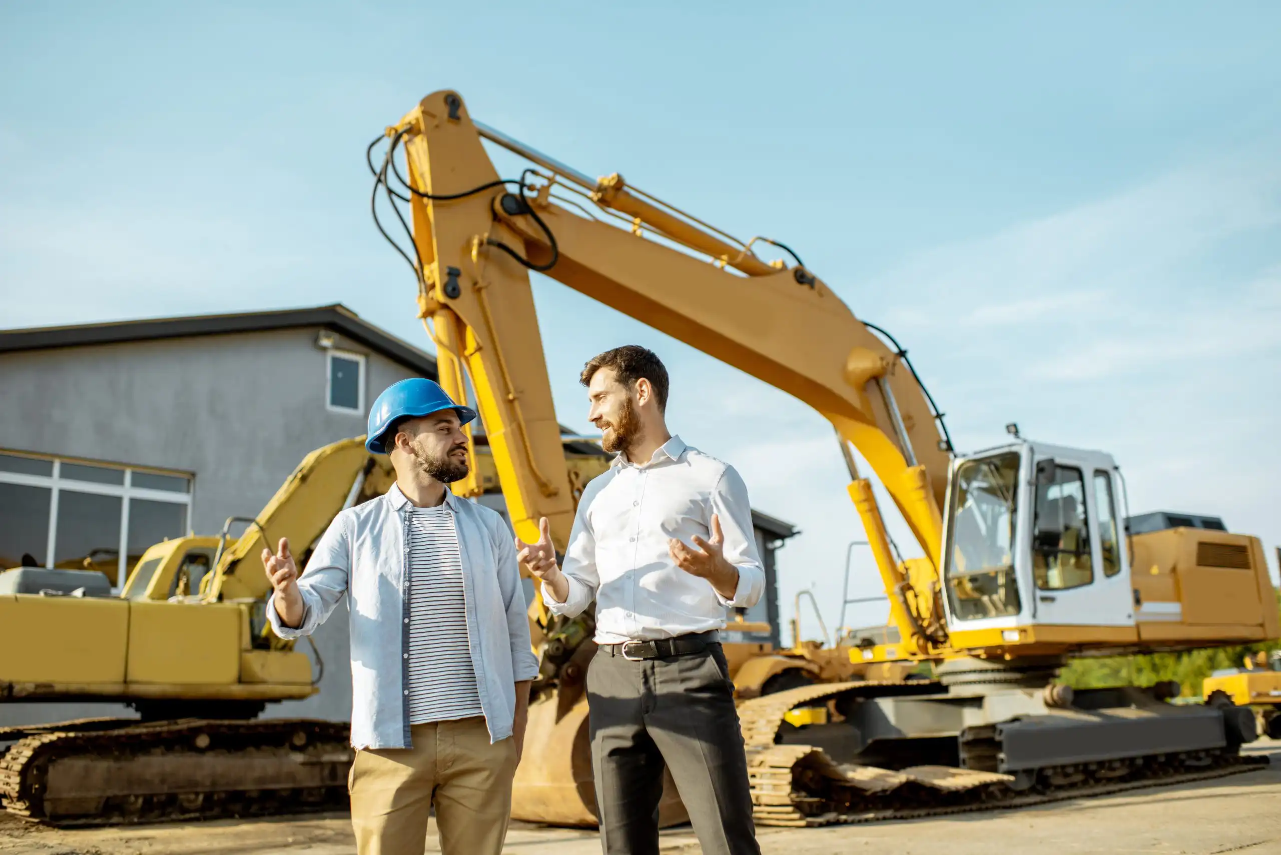 Two men in casual shirts stand and talk in front of a large yellow excavator on a construction site.