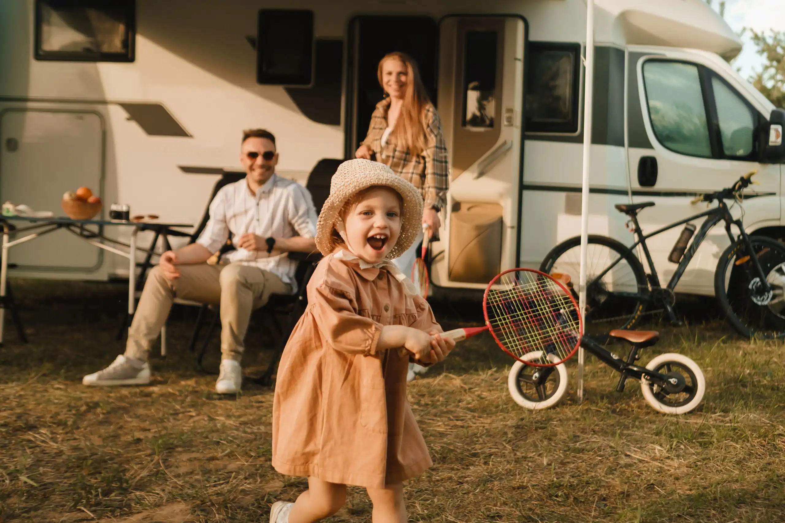 A young girl in a tan dress and straw hat plays with a red badminton racket in front of a camper van and family in the yard.