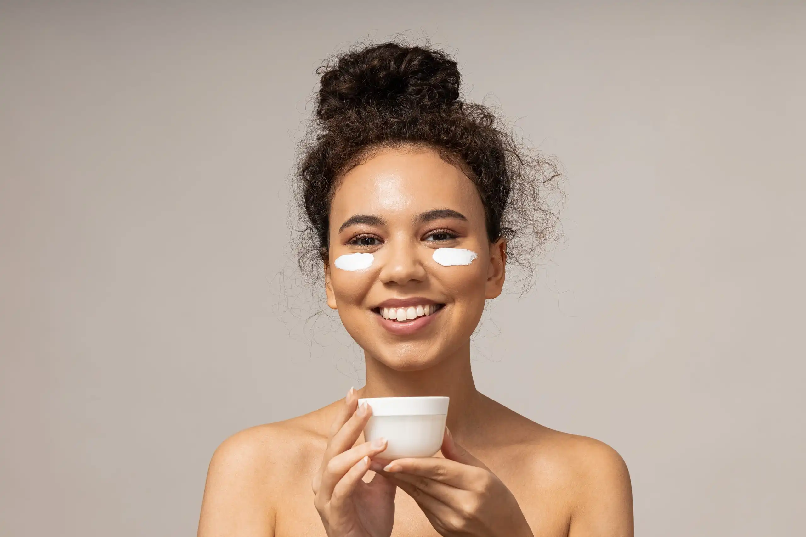 Smiling woman with white under-eye patches, holding a white cup against a neutral background.