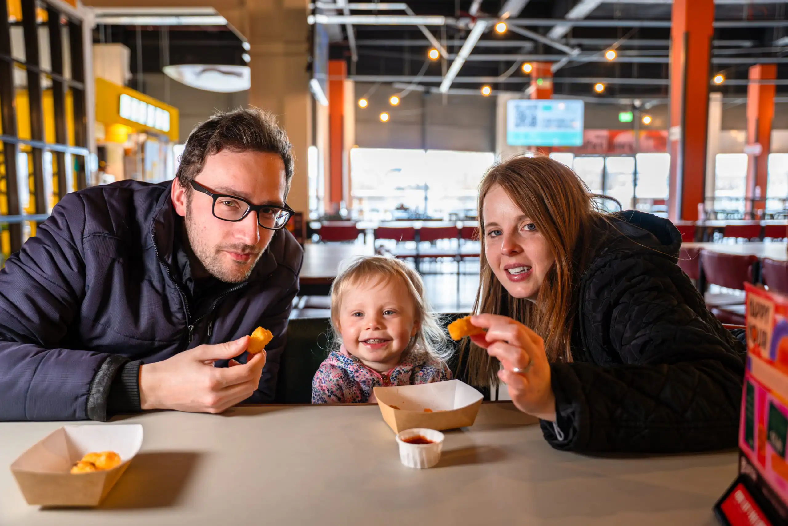 Family of three eating snacks at a casual restaurant, man on left, woman on right, smiling toddler in center.