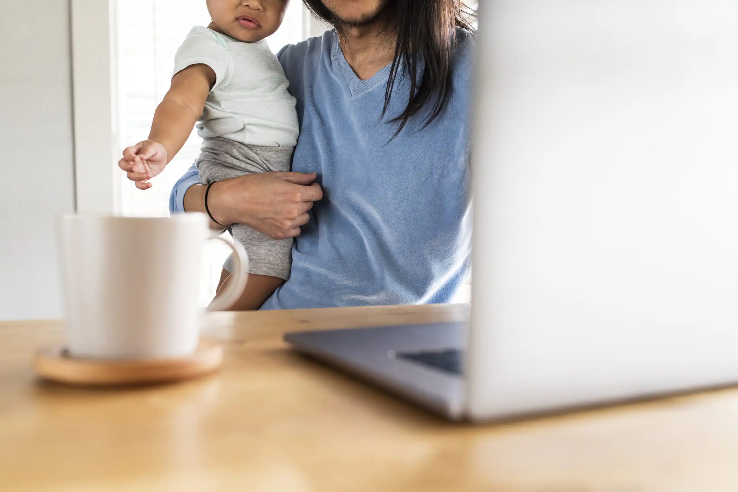 Parent holding a toddler at a bright kitchen table, with a mug and laptop visible in the foreground.