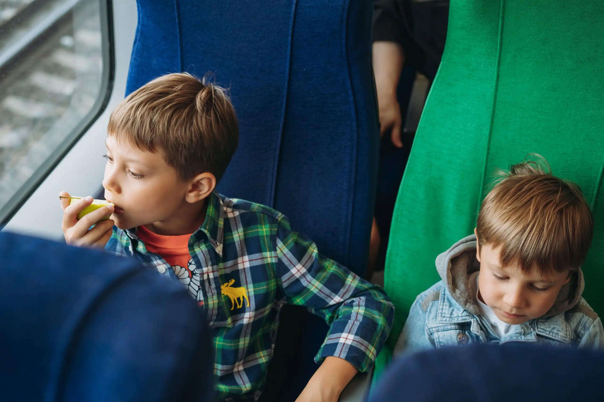children travelling by train. Boys sitting in train car and looking through window Two young children seated on a bus beside blue and green seats, looking down toward their hands or snacks.