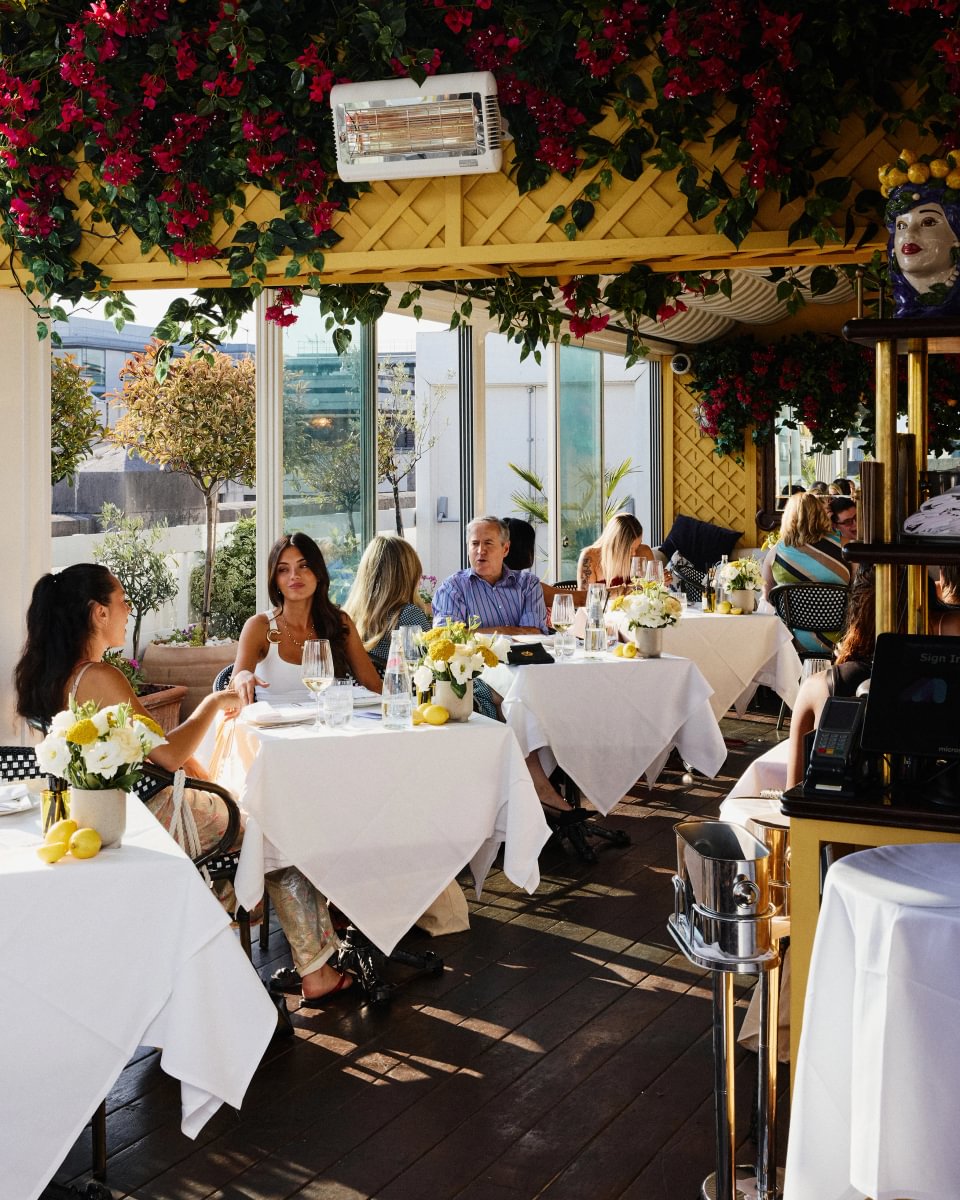 People dining on an outdoor terrace with white-tablecloth tables and pink flowers overhead.