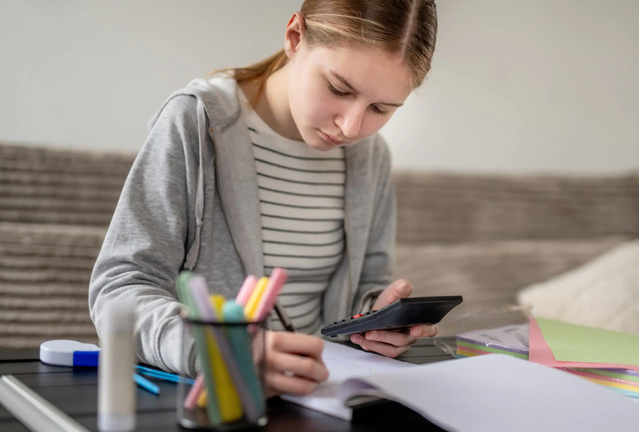 Girl in a gray hoodie writes in a lined notebook with colorful markers on a desk outdoors, focused on her work.