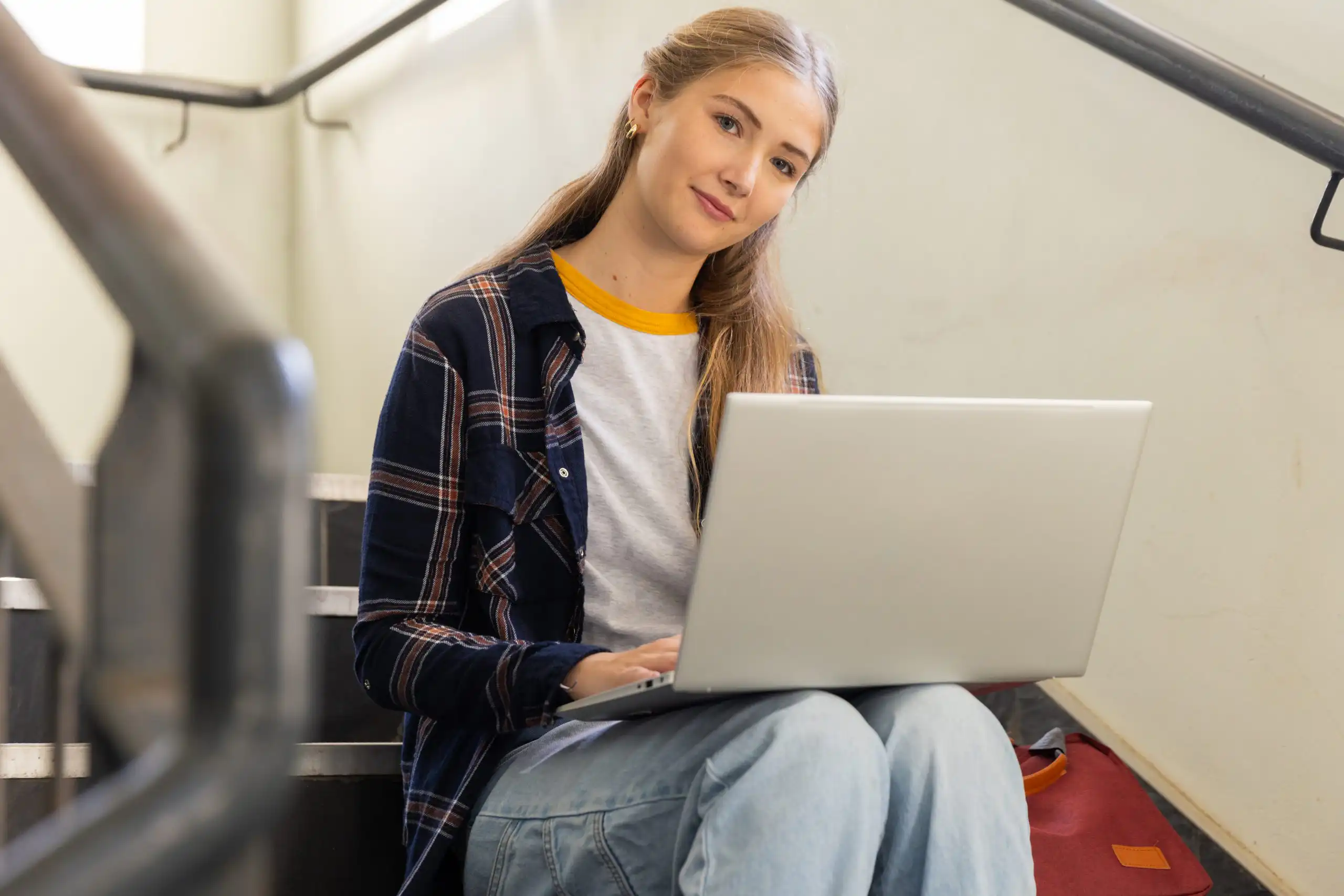 Young woman sitting on stairs with a laptop, wearing a plaid shirt and light jeans, looking at the camera with a slight smile.