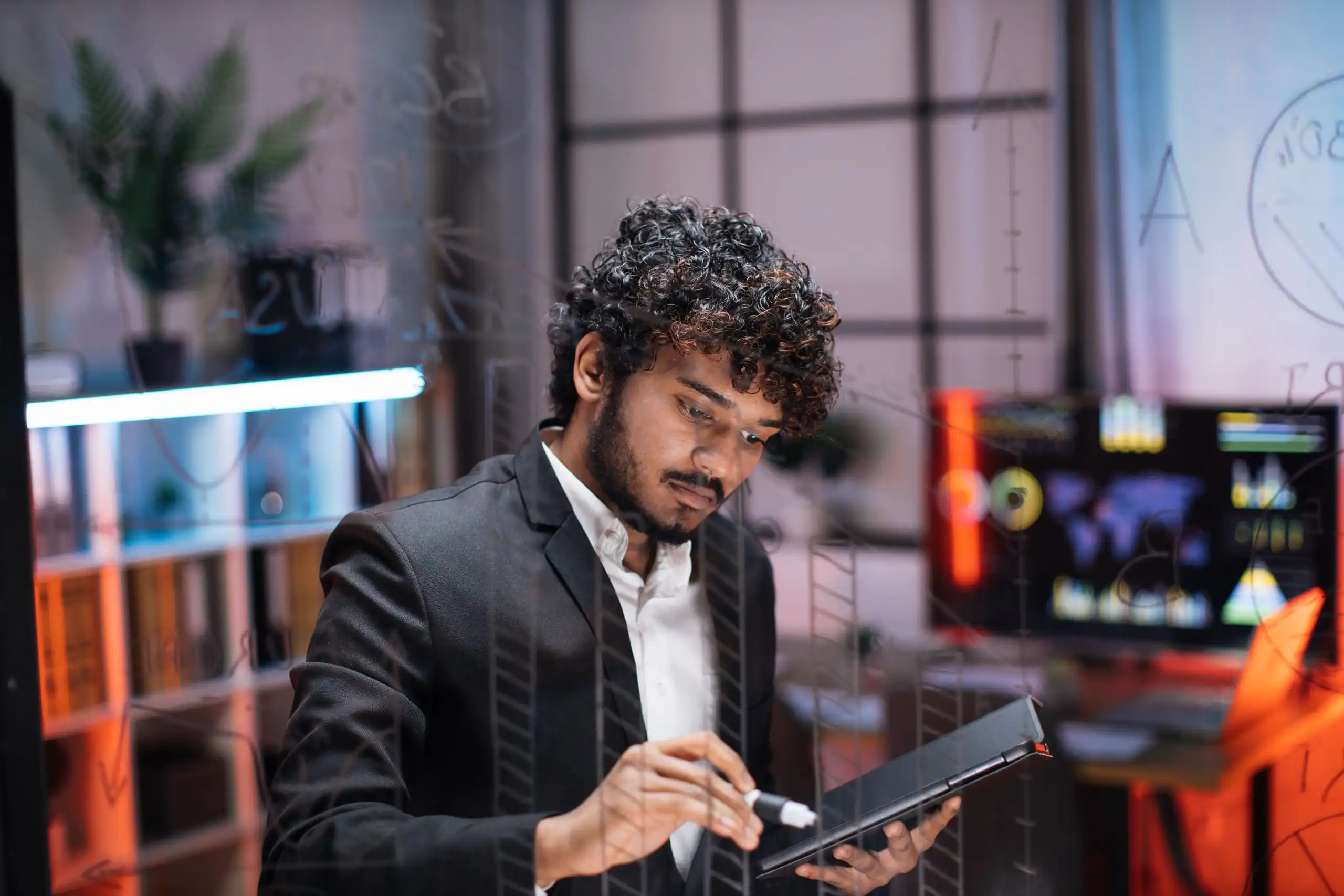 Man in a dark suit using a digital tablet at a desk in a modern glass-walled office setting with blurred screens in the background.