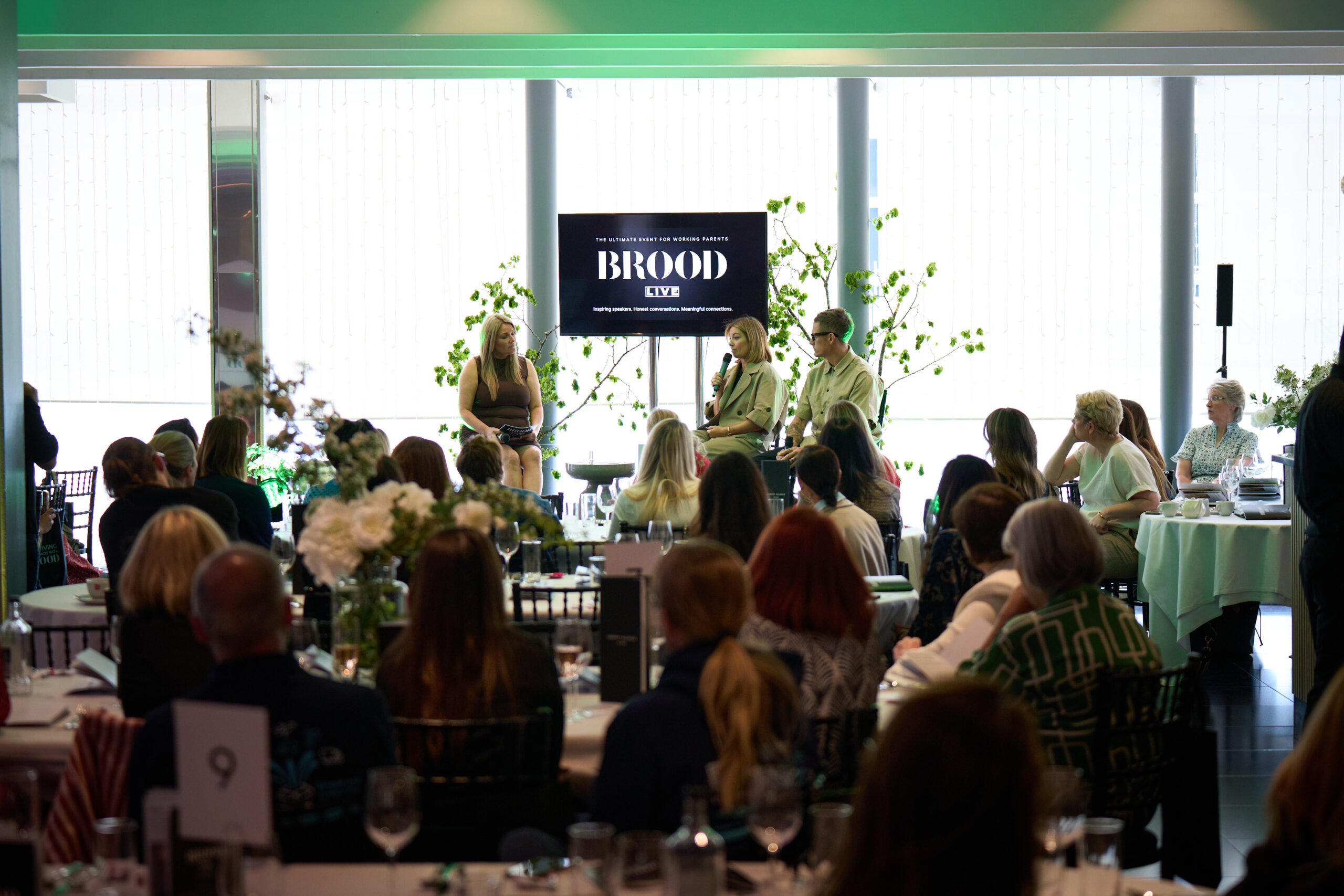 Three speakers in conversation on stage at a BROOD Live event, with a BROOD Live screen behind them and an audience seated at tables